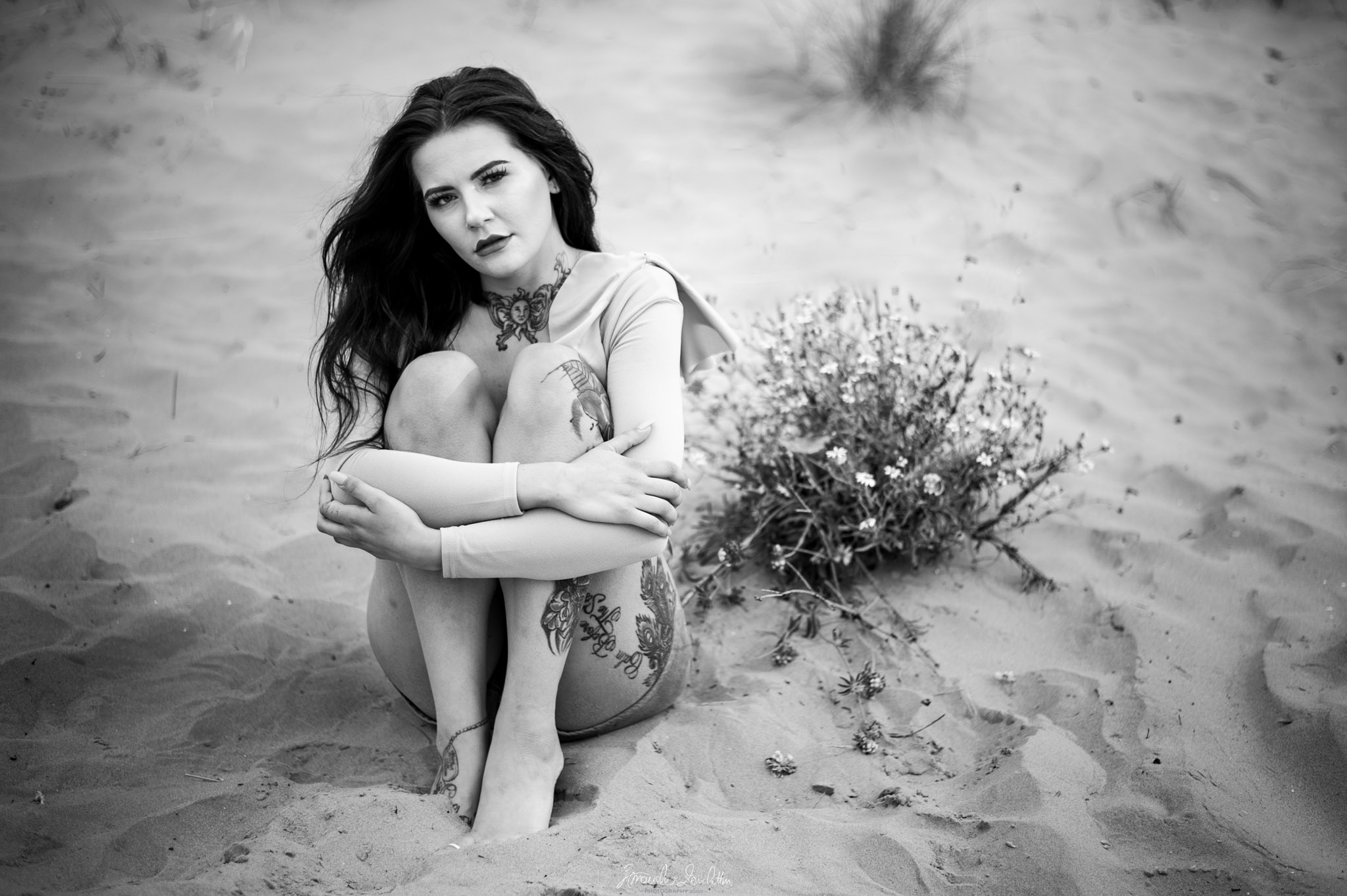 Black-and-white portrait of a woman seated on sand beside grasses